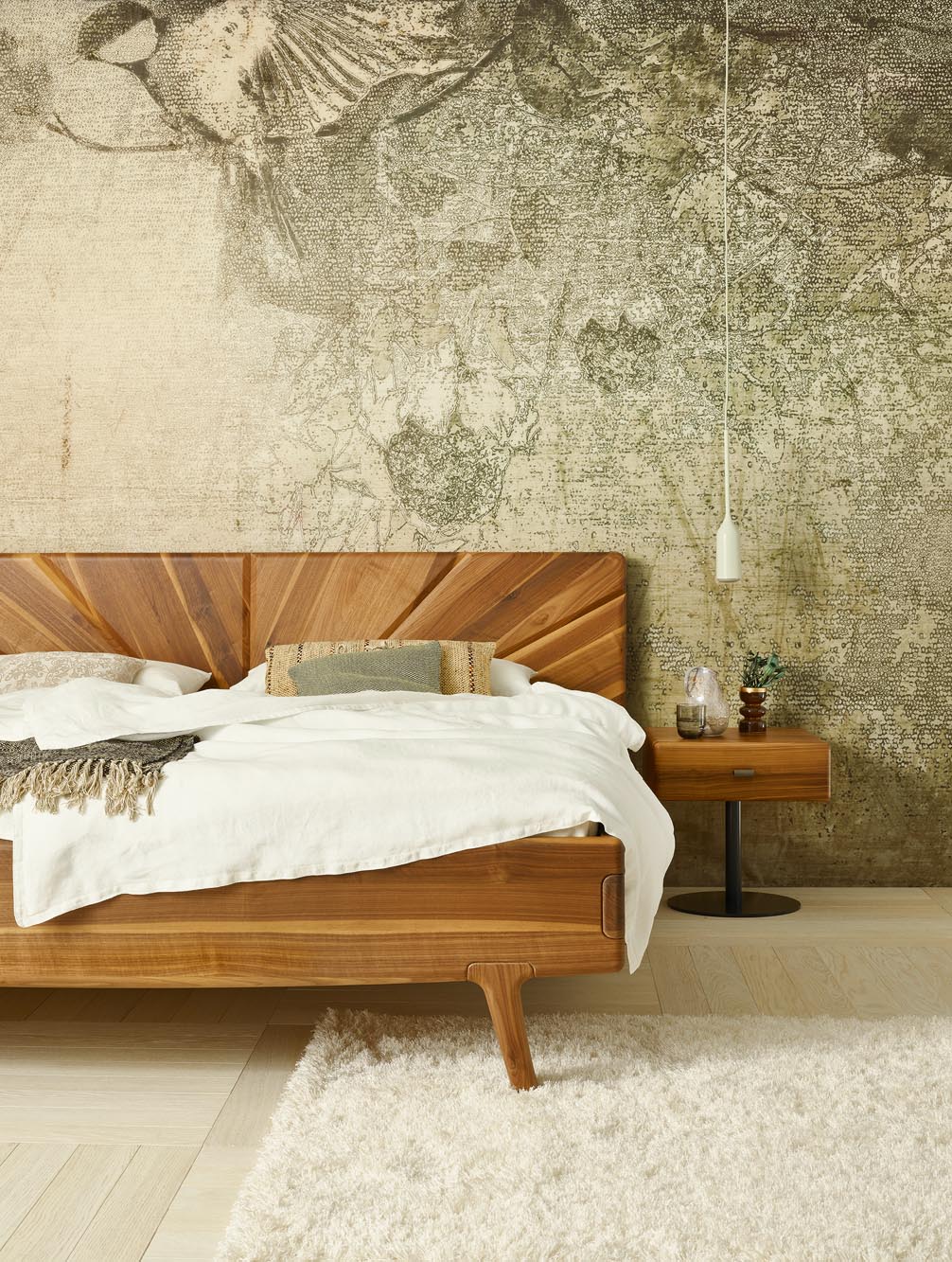 Ein modernes Schlafzimmer mit einem Anrei-Bettgestell aus Holz, weißer Bettwäsche, einer grauen Tagesdecke, einem Nachttisch aus Holz mit einer Pflanze und einer Lampe sowie einer strukturierten Wand in neutralen Farbtönen. Über dem Nachttisch hängt eine weiße Pendelleuchte.