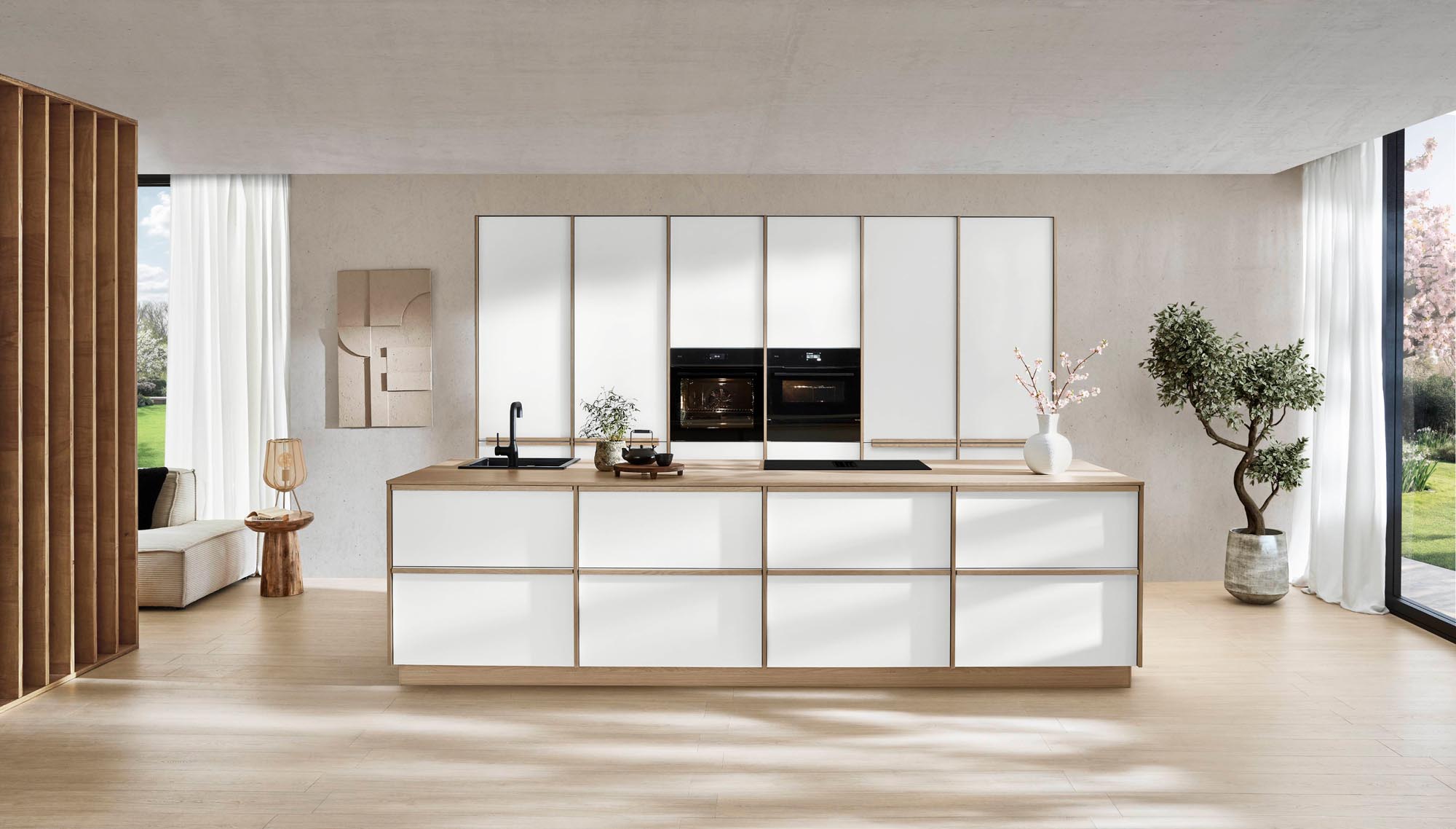 Minimalistische Küche mit weißen Schränken, eingebauten schwarzen Backöfen, einer zentralen Kücheninsel mit Spüle, Topfpflanzen, großen Fenstern und viel Tageslicht, das den Raum durchflutet.