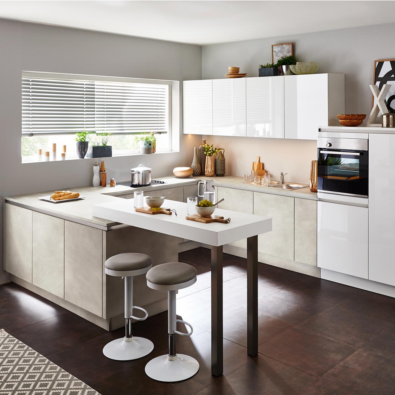 Moderne Küche mit weißen Schränken, einem Einbaubackofen, einem Herd, einem großen Fenster mit Jalousien und einer Frühstückstheke mit zwei Hockern. Auf den Arbeitsplatten stehen Lebensmittel, Geschirr und Küchenutensilien. Der Boden ist dunkel, und der Raum wirkt hell und sauber.