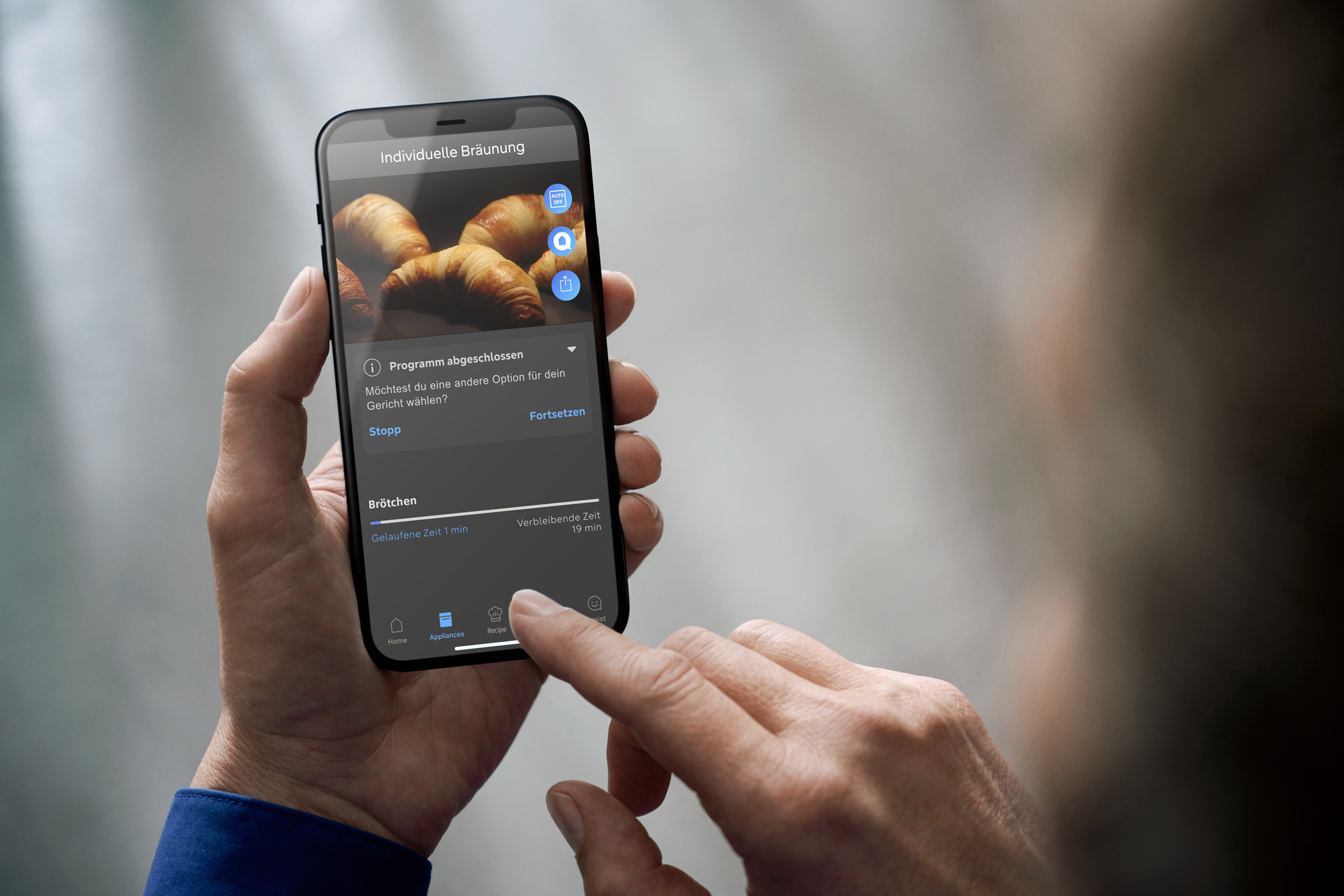 Eine Person hält ein Smartphone in der Hand, auf dem eine Siemens-App zum Thema Essen in deutscher Sprache angezeigt wird, die ein Bild von Croissants und verschiedene Einstellungen zeigt. Die Person bedient den Touchscreen mit dem Daumen.