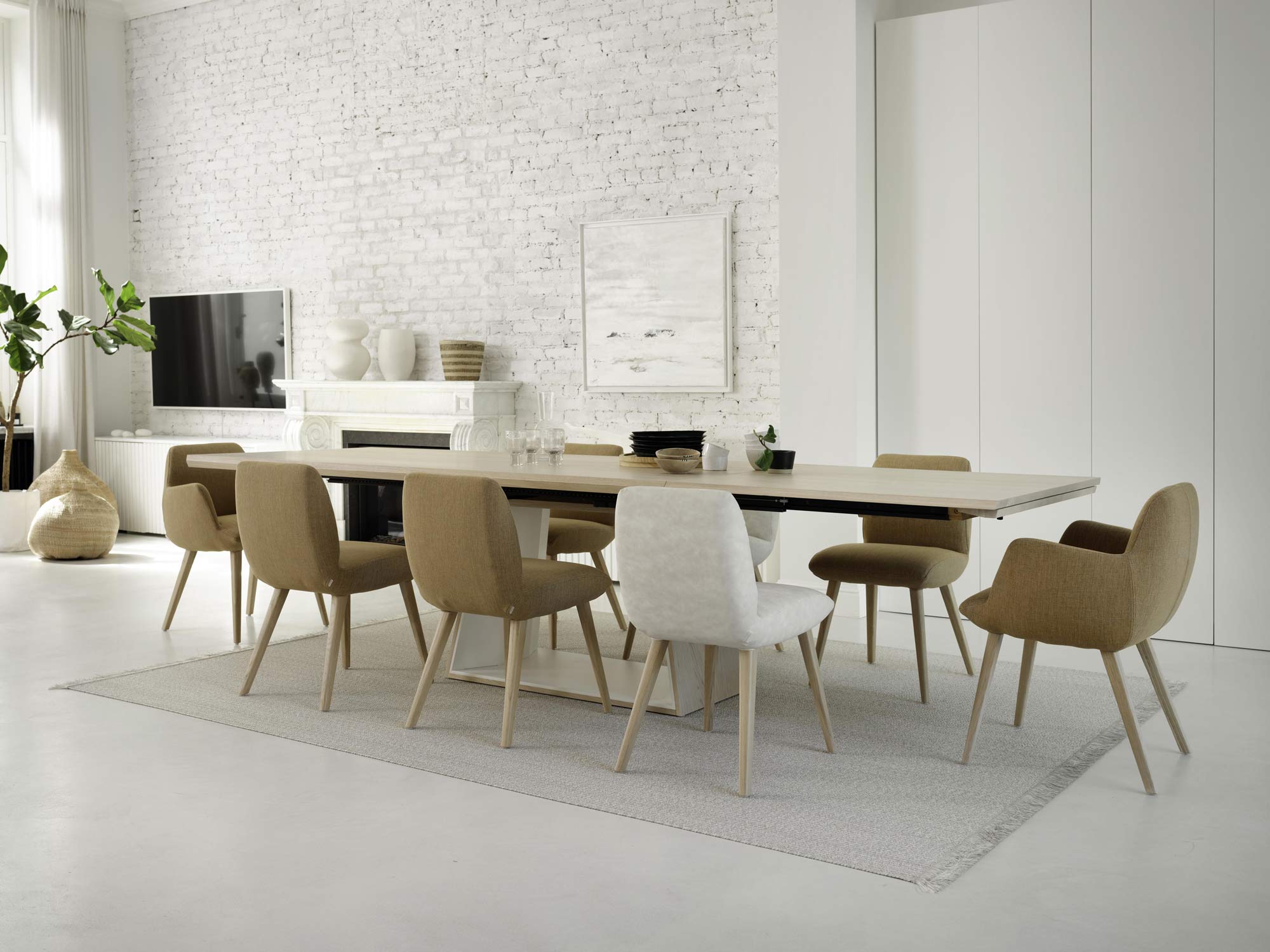 Ein modernes Esszimmer mit einem langen ausziehbaren Tisch, um den herum auf einem hellen Teppich verschiedene beige und weiße Stühle stehen. Der Raum besticht durch weiße Ziegelwände, eine minimalistische Einrichtung, große Fenster und eine Topfpflanze in der Ecke.