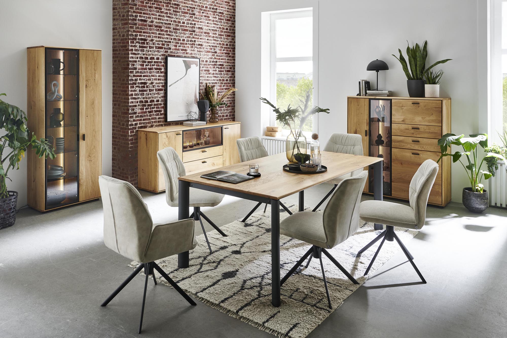 Modernes Niehoff-Esszimmer mit einem Tisch aus hellem Holz, sechs grauen Stühlen mit Polsterung, passenden Holzschränken, grünen Pflanzen, großen Fenstern, einer Akzentwand aus Ziegelsteinen und dekorativen Akzenten auf einem gemusterten Teppich.