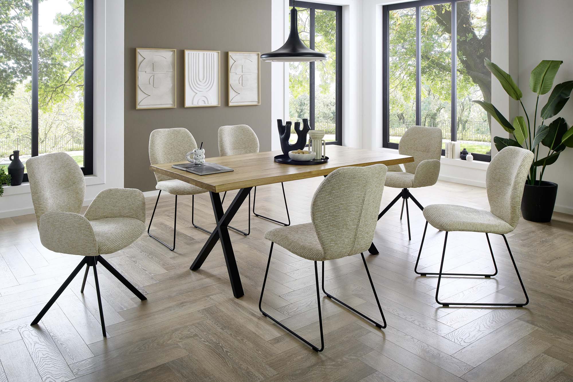 Modernes Esszimmer mit einem Holztisch von Niehoff, sechs cremefarbenen Stühlen, einer Hängeleuchte, abstrakter Wandkunst, großen Fenstern mit Blick ins Grüne und einer Topfpflanze in der Ecke.