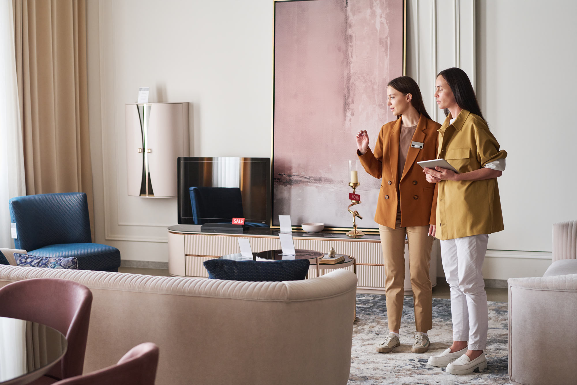 Zwei Frauen stehen in einem modernen, stilvollen Wohnzimmer mit neutralen Möbeln von Sudbrock, abstrakten Kunstwerken und Dekorationsgegenständen. Die eine hält ein Tablet in der Hand, während die andere gestikuliert, während sie sich unterhalten oder durch den Raum gehen.