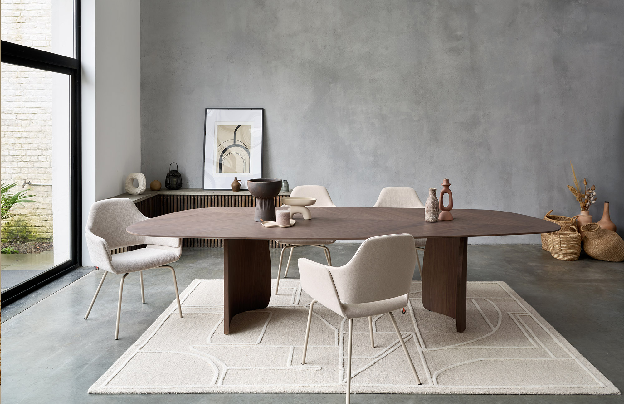 Ein modernes Esszimmer mit einem dunklen Holztisch, sechs hellen Polsterstühlen, einer neutralen Einrichtung, Kunstwerken auf einer Anrichte, geflochtenen Körben und einem großen Fenster, durch das Tageslicht hereinströmt.