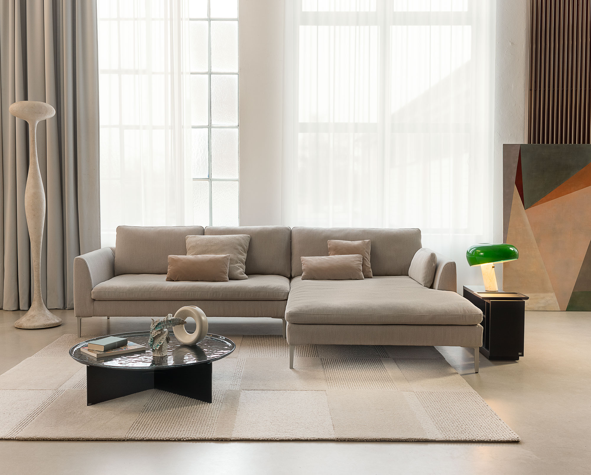 Ein modernes Wohnzimmer mit einem beigen Ecksofa, drei Kissen, einem runden schwarzen Couchtisch mit Büchern und Dekoartikeln, einer Stehlampe, einem Beistelltisch mit einer grünen Glaslampe und großen Fenstern mit transparenten Vorhängen.