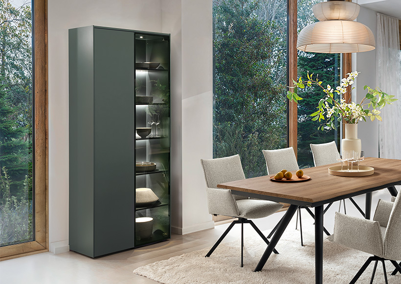 Modernes Esszimmer mit einem Holztisch von Venjakob, sechs hellen Stühlen, einer Glasvitrine mit Geschirr, großen Fenstern und einer Pendelleuchte. Durch die Fenster sind draußen grüne Bäume zu sehen.